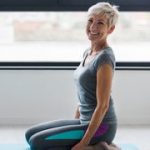 Dieses Bild zeigt eine Frau kniend auf einer Matte in einem Studio. Sie bereitet sich vor auf eine kommende Yoga-Session. Die Frau lächelt und ist zufrieden mit Ihrer Gesundheit.